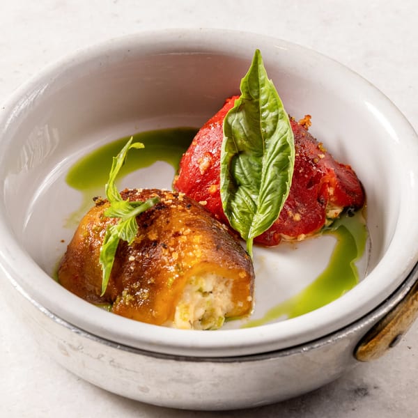 Two stuffed peppers, one red and one golden, garnished with basil and green sauce in a white bowl.