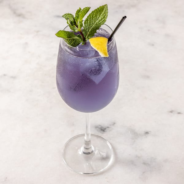 A purple cocktail in a wine glass, garnished with mint, a lemon wedge, and a black straw.