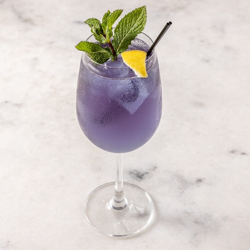 A purple cocktail in a wine glass, garnished with mint, a lemon wedge, and a black straw.