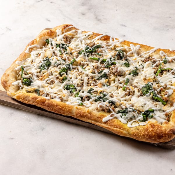 Rectangular Italian pizza with white sauce, crumbled sausage, and spinach on a wooden board.