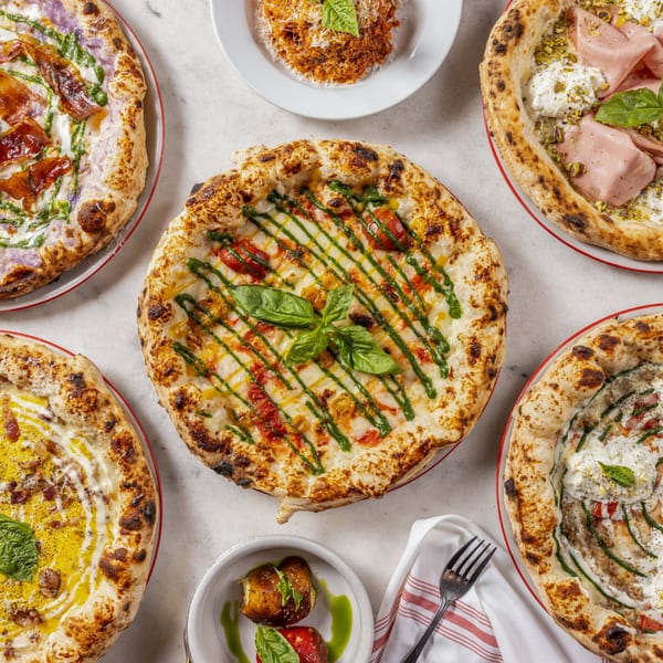 Five gourmet pizzas with various toppings, a pasta dish, and an appetizer on a white marble surface.