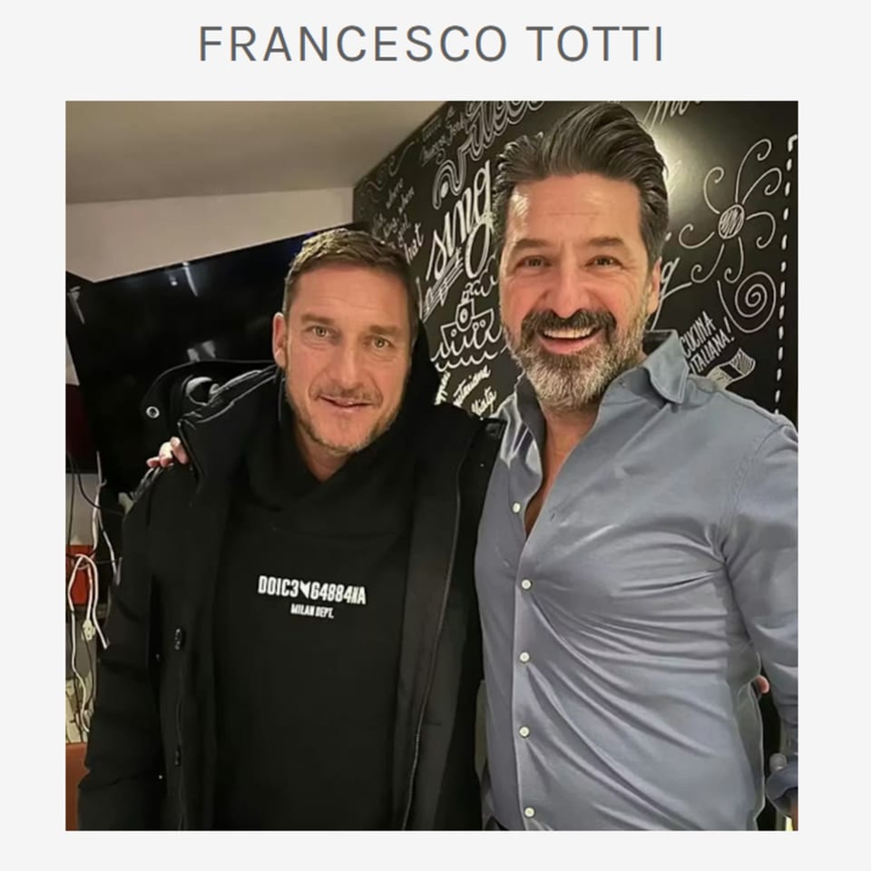 Two men, Francesco Totti and another, smiling at the camera in an Italian restaurant.