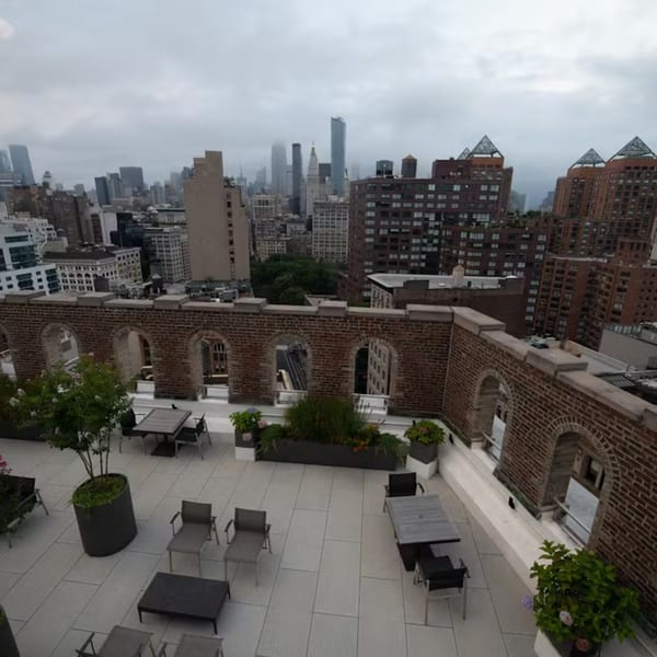 Rooftop patio with tables and chairs, surrounded by a brick wall with arched openings, overlooking a city skyline.