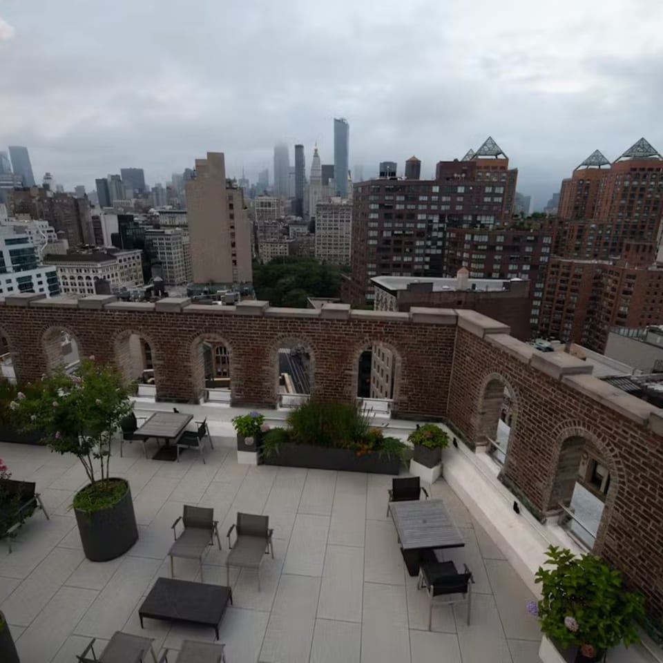Rooftop patio with tables and chairs, surrounded by a brick wall with arched openings, overlooking a city skyline.
